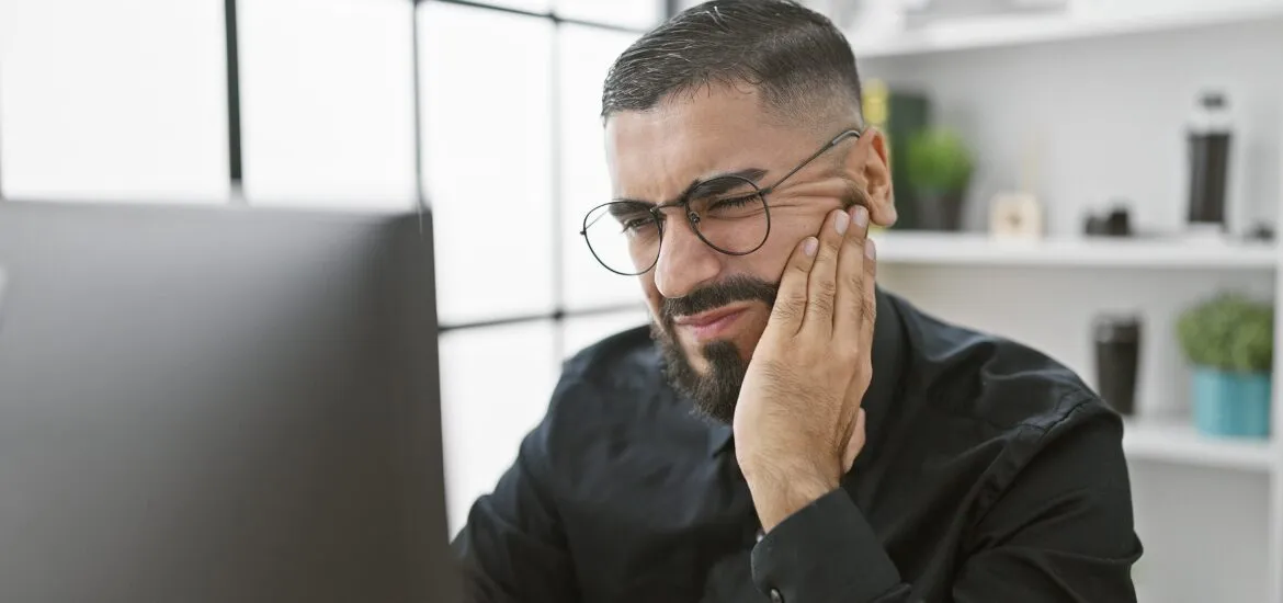 A young bearded man expresses toothache discomfort at his bright office workspace
